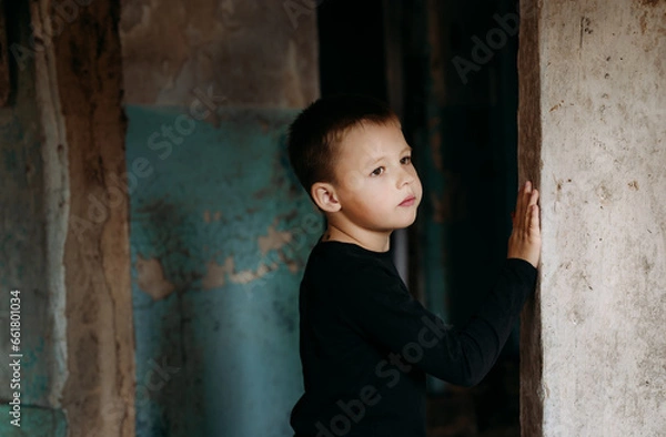 Fototapeta Close-up of a boy in a black t-shirt standing by the door and looking out the window from his dilapidated house. War in Ukraine. Consequences of war.