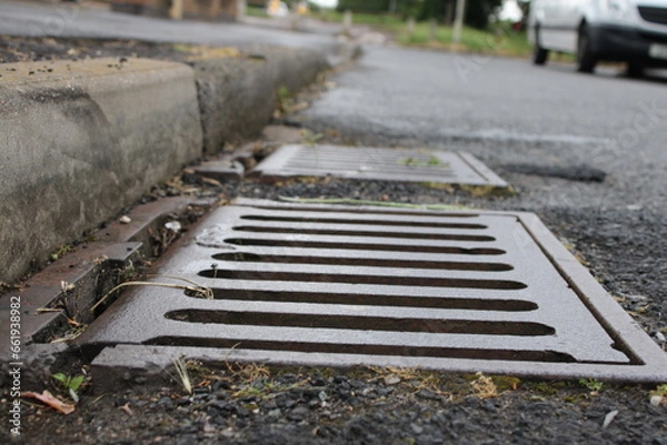 Fototapeta Grate of a storm sewer drain up close perspective, blurred street in background