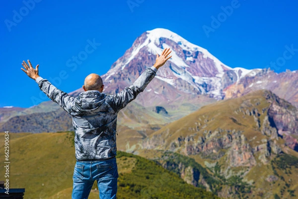 Fototapeta the traveler, raising his hands up, enjoys the picturesque view of the snow-capped peak of Mount Kazbek. Travel in Georgia