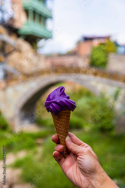 Fototapeta A hand holds a wine ice cream cone on a blurred background of the old city of Tbilisi. Wine ice cream in a waffle cone.