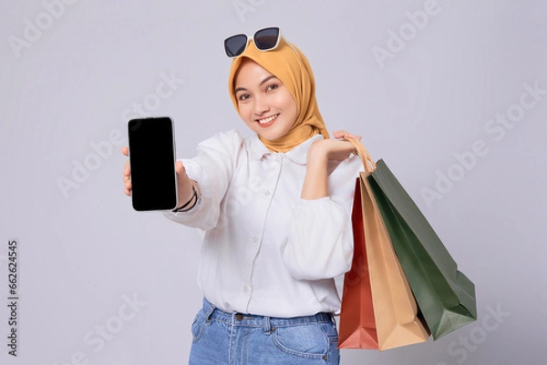 Fototapeta Beautiful smiling young Asian Muslim Woman showing mobile phone with a blank screen and holding shopping bags isolated on white background. online mobile shopping concept