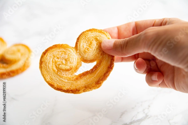 Obraz Young woman's hand holding a puff pastry palmier on a light background. Homemade bakery.