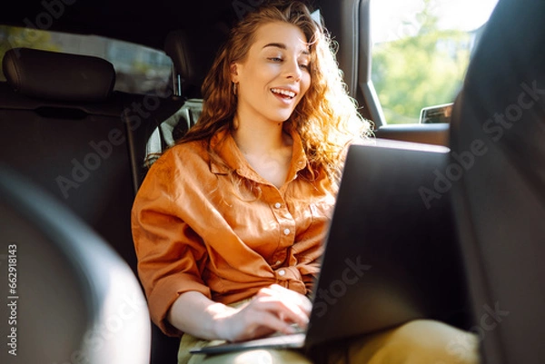 Fototapeta A young curly woman with a laptop sitting in the back seat of a car. Beautiful woman uses laptop in car. Remote work, freelance.