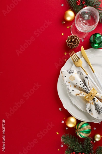Fototapeta Transform your space: the road to a beautiful Christmas table. Top view vertical shot of cutlery, plates, glassware, fir twigs, tree ornaments, cones, stars confetti on red background with ad spot