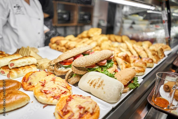 Fototapeta Fresh sandwiches, pies and pizza to choose from in the window of a cafe, buffet or self-service cafe. Croissant with ham and cheese. Hot pastries lie on the shelf in the cafe. Buns in the store.