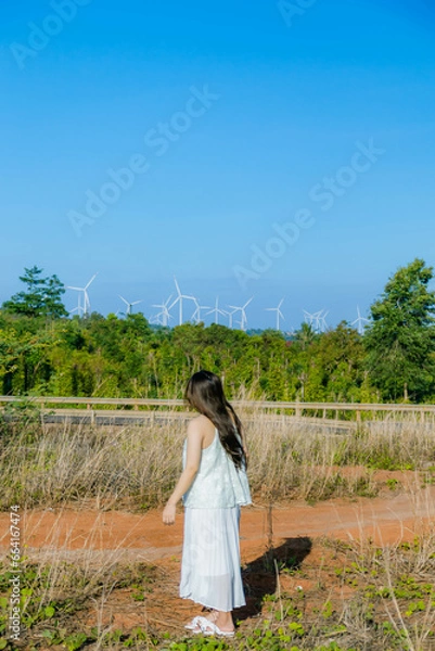 Fototapeta A Girl stands against the wind turbine generate electricity, fan fields on the green grass over the blue clouded sky in Ea H'leo district, Dak Lak province, Vietnam.