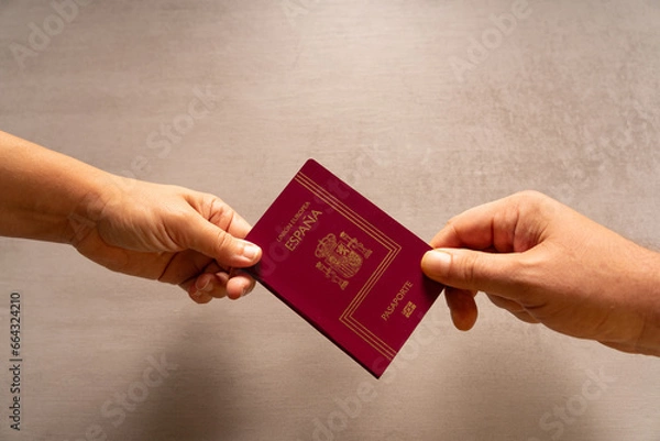 Obraz A hand of a man and another of a woman hold a passport. On a table in the background