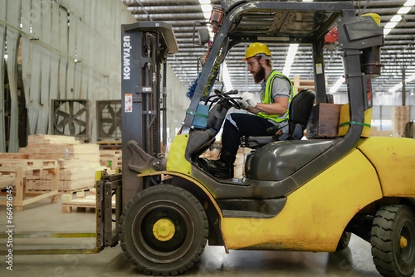 Fototapeta Warehouse worker wears safety helmet driving forklift truck in pallet factory. Skilled male logistic engineer working in shipping storage manufacturing lifting, moving and unloading cargo for delivery