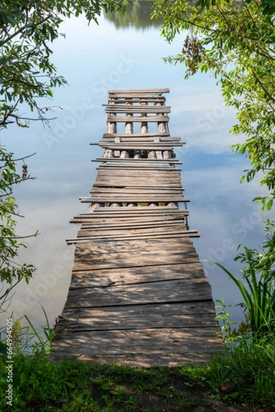 Fototapeta A wooden bridge made of logs and partially rotten boards. Being careful is dangerous. Water, lake, bush. Shore.