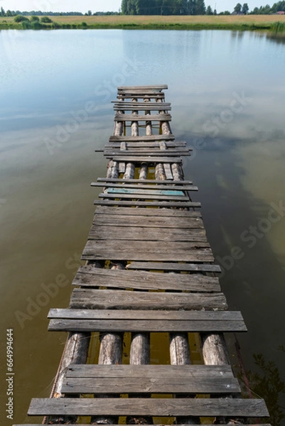 Fototapeta A wooden bridge made of logs and partially rotten boards. Being careful is dangerous. Water, lake, bush. Shore.