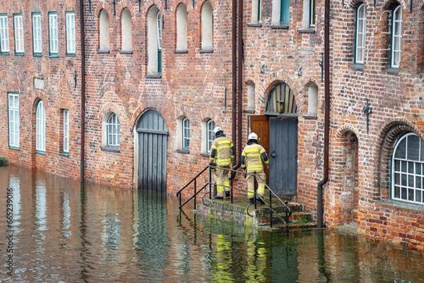 Fototapeta High water flooding the old town of Lubeck when the river Trave overflows its banks, specialists control water level and danger at a historic building of red brick architecture