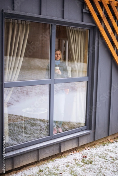 Obraz Girl looking through window with hot drink cup in both hands, snow outside and warm inside, ppond and snow reflection can be seen on the glass