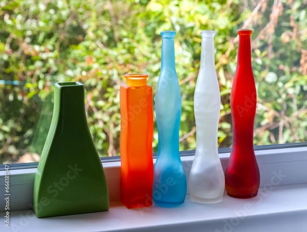 Obraz Multicolored glass decorative bottles on a white window sill against a background of greenery