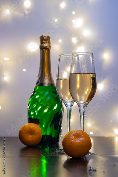 Obraz Crystal champagne glasses, tangerines, a bottle of champagne and garland lights in the background. Champagne, tangerines and garlands create a festive mood.