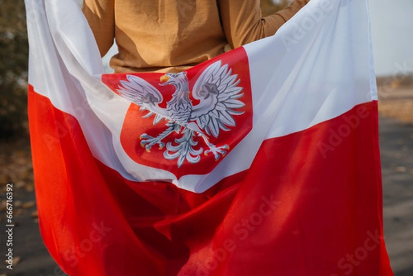 Fototapeta A little boy behind his back is holding the flag of Poland in his hands. Child standing on road with national flag on nature. Close-up. Concept of national holidays and patriotic events. Patriotism
