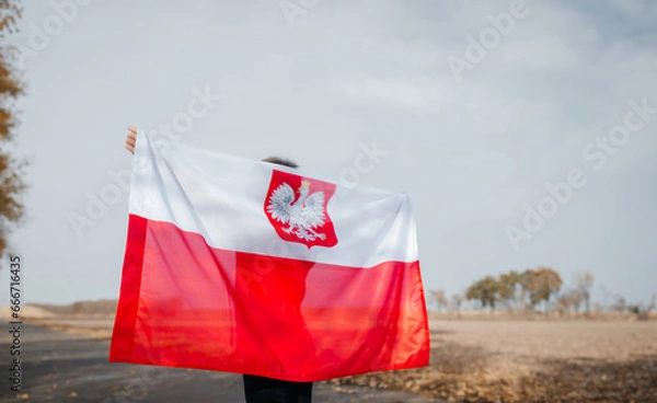 Fototapeta A little boy behind his back is holding the flag of Poland in his hands. Child standing on road with national flag on nature. Close-up. Concept of national holidays and patriotic events. Patriotism