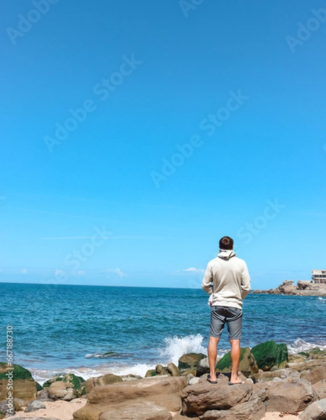 Fototapeta Young man traveler contemplating in a peaceful place in fornt of the ocean viewed from the back. Portugal Solo traveler.