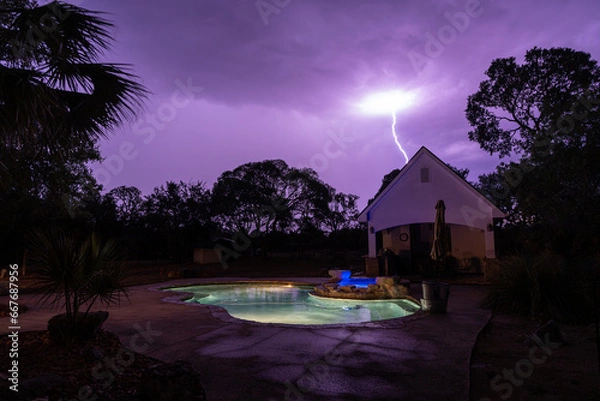 Obraz A night time lightning strike a backyard structure with a pool, a spa and trees under a purple dark cloudy sky, Hill Country, Texas