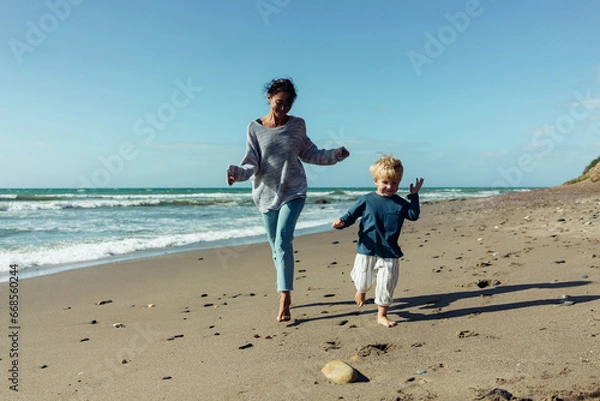 Obraz A young mother and her three-year-old son run, play, smile, and laugh on the seashore.