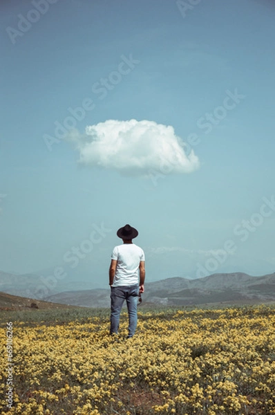 Fototapeta A boy with a white t-shirt and a hat on his head and glasses in his hand with a special and single cloud in the sky in a field full of yellow flowers