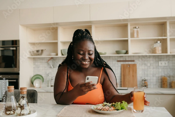 Obraz Beautiful plus size African woman using smart phone while enjoying healthy eating for lunch at home
