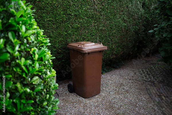 Fototapeta Brown large plastic bin for trash on backyard, around of tree hedge.