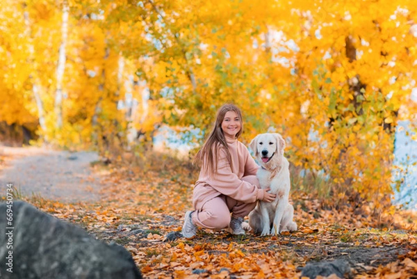 Obraz A girl in an autumn park walks with a golden retriever dog. There are a lot of fallen autumn leaves around. A river flows in the background.