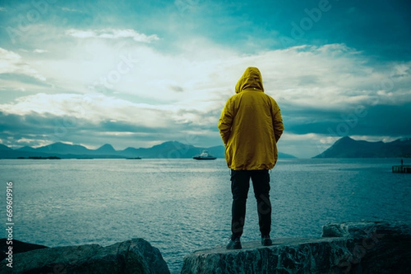 Fototapeta Guy standing on the rock and admiring a beautiful scenery of Norway fjord and mountains. Rainy weather and warm clothing.