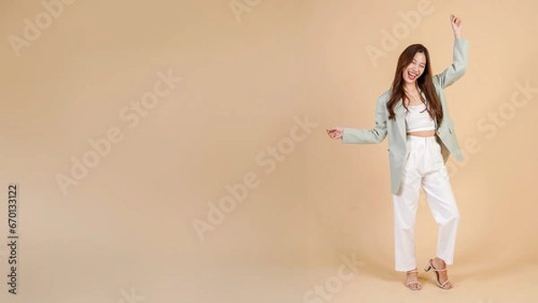 Obraz Young Asian happy smiling woman in casual business suit raising her hands up like a winner isolated over beige background - with empty copy space - in full body