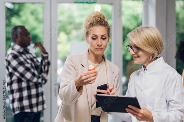 Fototapeta Senior pharmacist talking with a customer in a pharmacy and using tablet