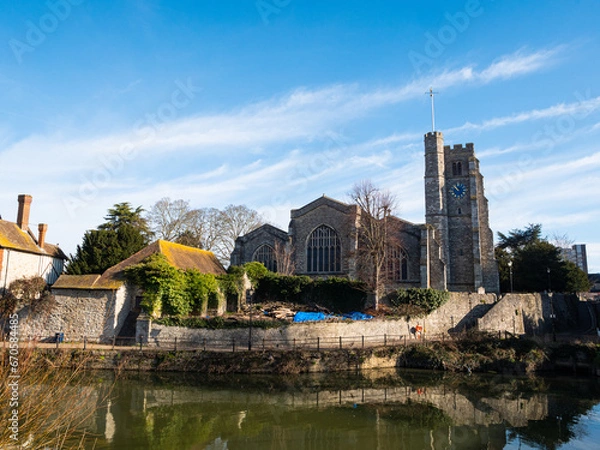 Fototapeta In foreground, the River Medway flows past All Saints Church, a paved riverside walkway and period stone houses at Maidstone, Kent. Bright blue sky, light clouds.