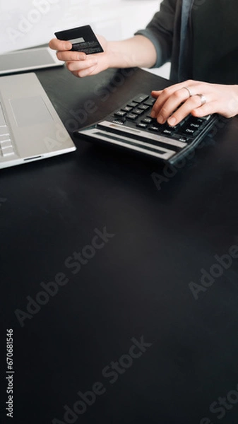 Fototapeta Woman accountant in office, using calculator and holding credit card. Close-up of hands. Online shopping and finance concept. Financial management, digital transactions, business and finance.