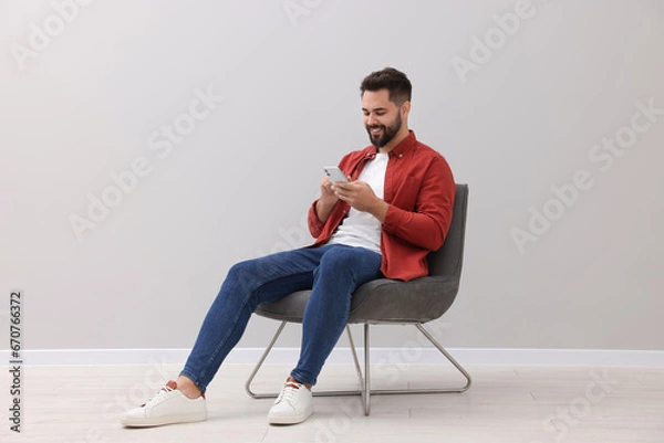 Fototapeta Handsome man using smartphone while sitting in armchair near light grey wall indoors