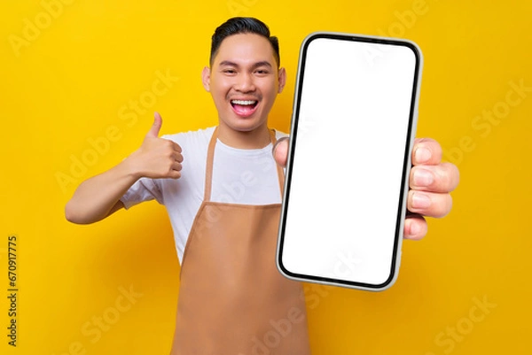 Fototapeta excited young Asian man barista barman employee wearing brown apron work in coffee shop holding mobile phone and showing thumb up sign isolated on yellow background. Small business startup concept