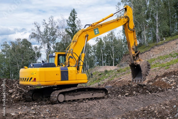 Fototapeta A yellow excavator works in the mountains. Construction of roads and houses in beautiful mountains.