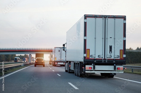 Fototapeta Modern white semi-trailer trucks on the highway driving in the right lane. Commercial vehicle for shipping and post delivery. Shipping of the goods on land with a door-to-door delivery process.