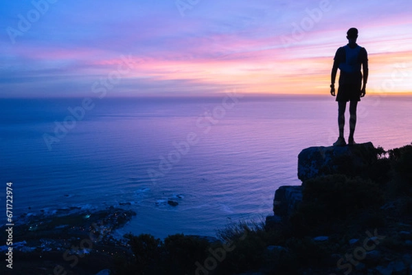 Fototapeta A hiker silhouetted against the sunset over the ocean while standing on the top of a mountain that they have successfully climbed.