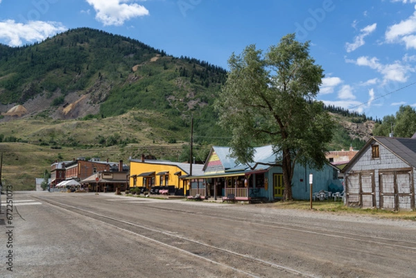 Fototapeta A strip of old style wooden and brick buildings along an old town street with trees and mountains in the background, Silverton, Colorado