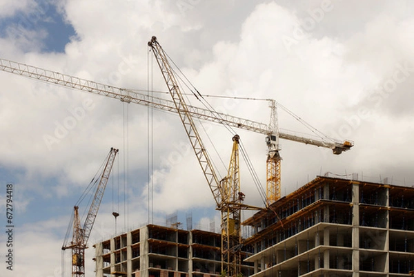 Fototapeta Tower cranes work during the construction of a multi-story building. New apartments for residents and premises for offices. Risky work at height. Lifting heavy building materials. City development
