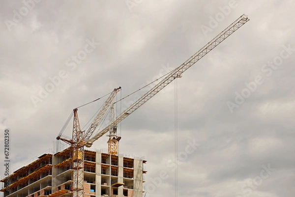 Fototapeta Tower cranes work during the construction of a multi-story building. New apartments for residents and premises for offices. Risky work at height. Lifting heavy building materials. City development