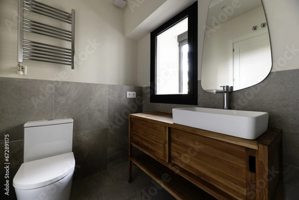 Fototapeta Small modern design bathroom with gray stone effect tiles, mirror on the wall, white porcelain sink cantilevered on wooden cabinet, towel drying radiator and chrome taps, black aluminum window