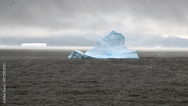 Obraz A slice of sunlight illuminates the flat Iceberg in the waters off Qaqortoq, Greenland, the town just visible in the haze to the right of the iceberg in the foreground.