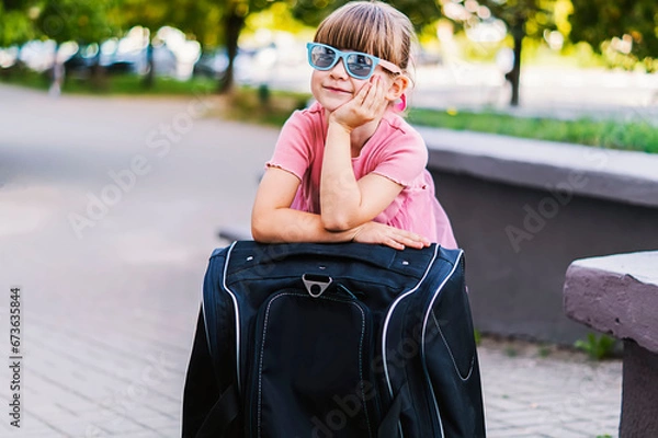 Obraz Close-up of a cute happy little traveler girl in sunglasses leaning on a black suitcase. Smiling pretty small girl with a suitcase. Children go abroad for summer holidays or weekends. Travel concept