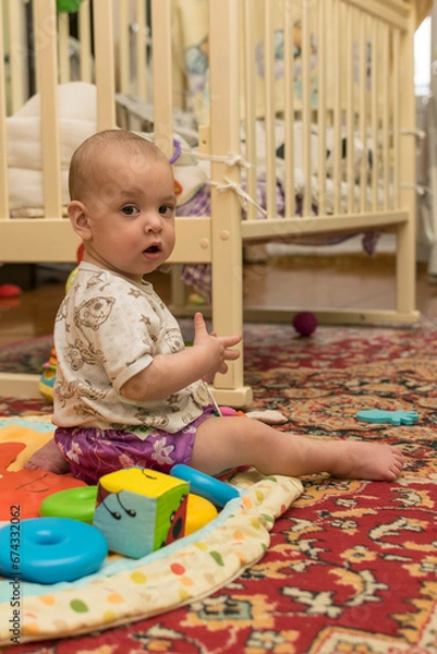 Fototapeta The baby is sitting on the floor near the crib and playing with toys.