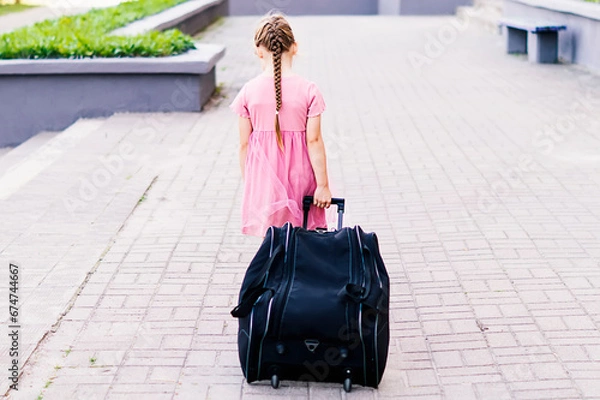 Obraz Rear view of a stylish little girl in a pink dress walking with luggage. Full length shot of a little girl walking and pulling a suitcase. A child rolls a suitcase on wheels along the sidewalk