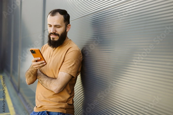 Obraz Caucasian bearded guy using smartphone leaning on a urban wall