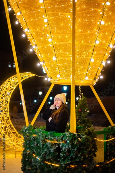 Fototapeta Beautiful woman in warm clothes, a hat and a coat in winter against the background of Christmas street lights. Celebrates the New Year on the street against the backdrop of street decor. bokeh