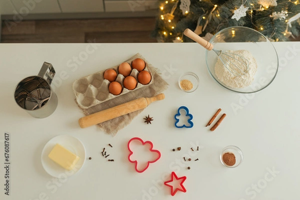 Fototapeta On the kitchen table there is a bowl with flour, a whisk, spices and molds for making Christmas cookies, top view
