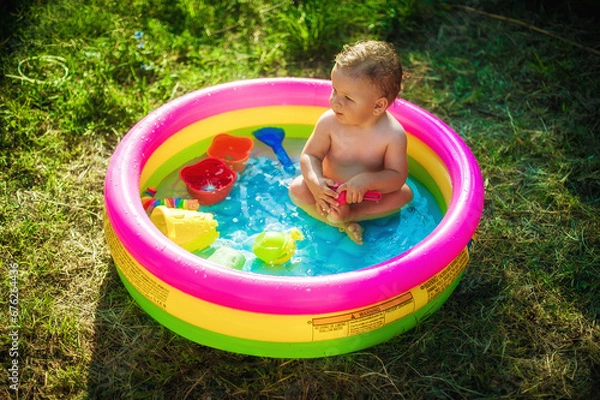 Fototapeta A cute little baby boy is playing with toys while bathing in a little colorful wading pool on meadow garden field outdoor. Summer time. Joy and happiness. Love and family