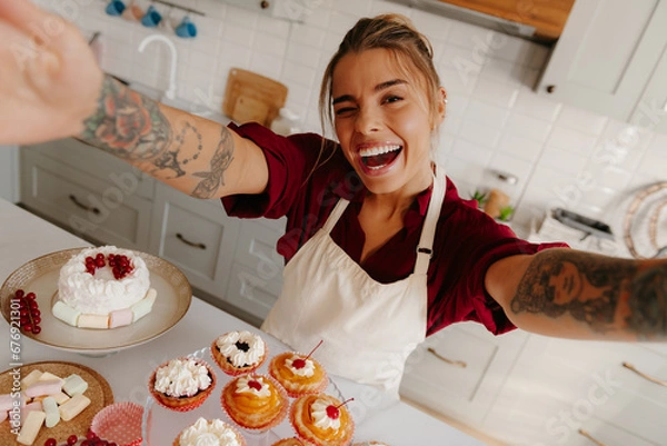Obraz Top view of playful female baker making selfie with variety of sweet food laying on kitchen table